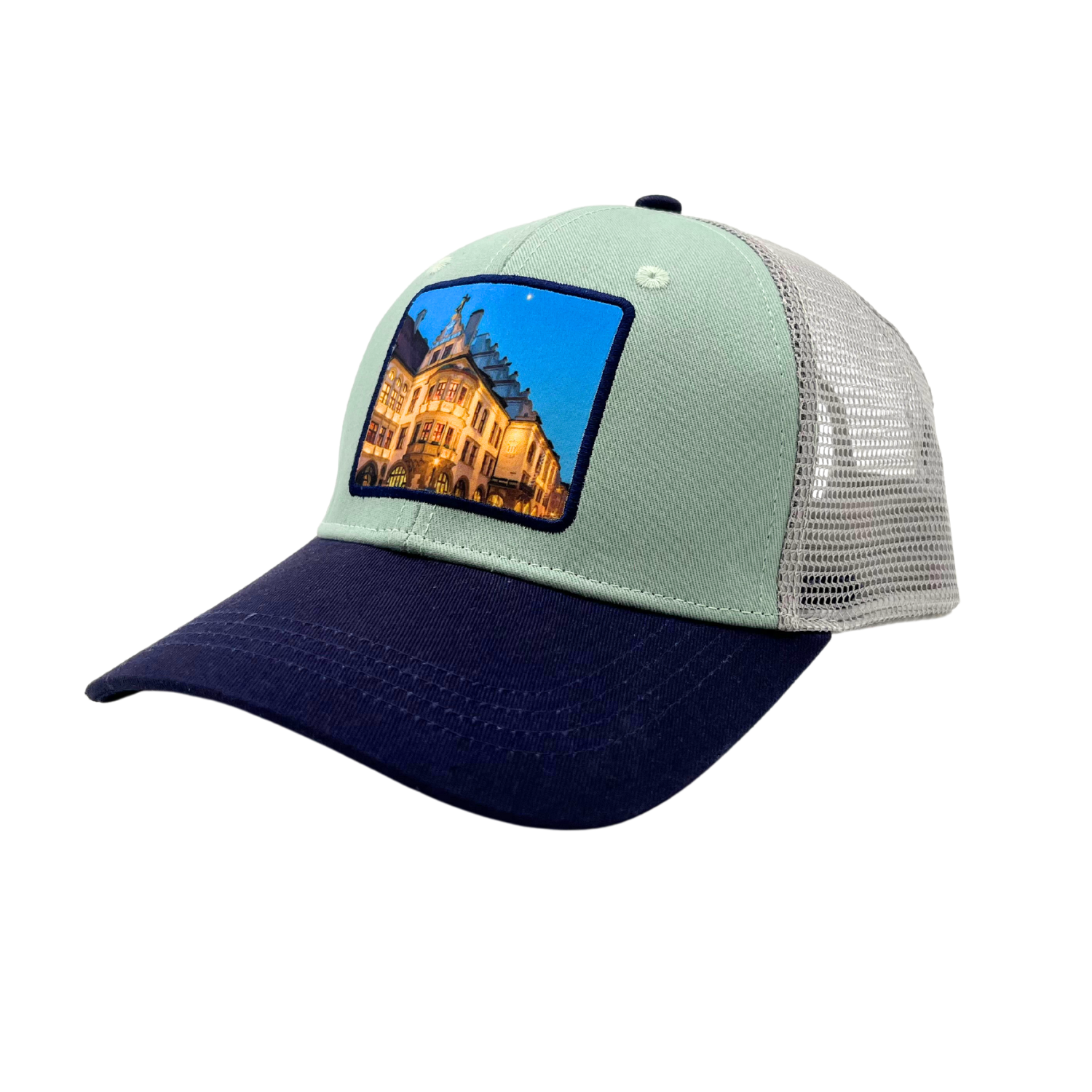 „Trucker-Cap mit hellgrauer Front, dunkelblauem Schirm und weißen Mesh-Einsätzen an den Seiten und hinten. Auf der Vorderseite befindet sich ein rechteckiger Aufnäher oder Fotodruck mit einem Bild des beleuchteten Hofbräuhauses vor sternenklarem Himmel.“