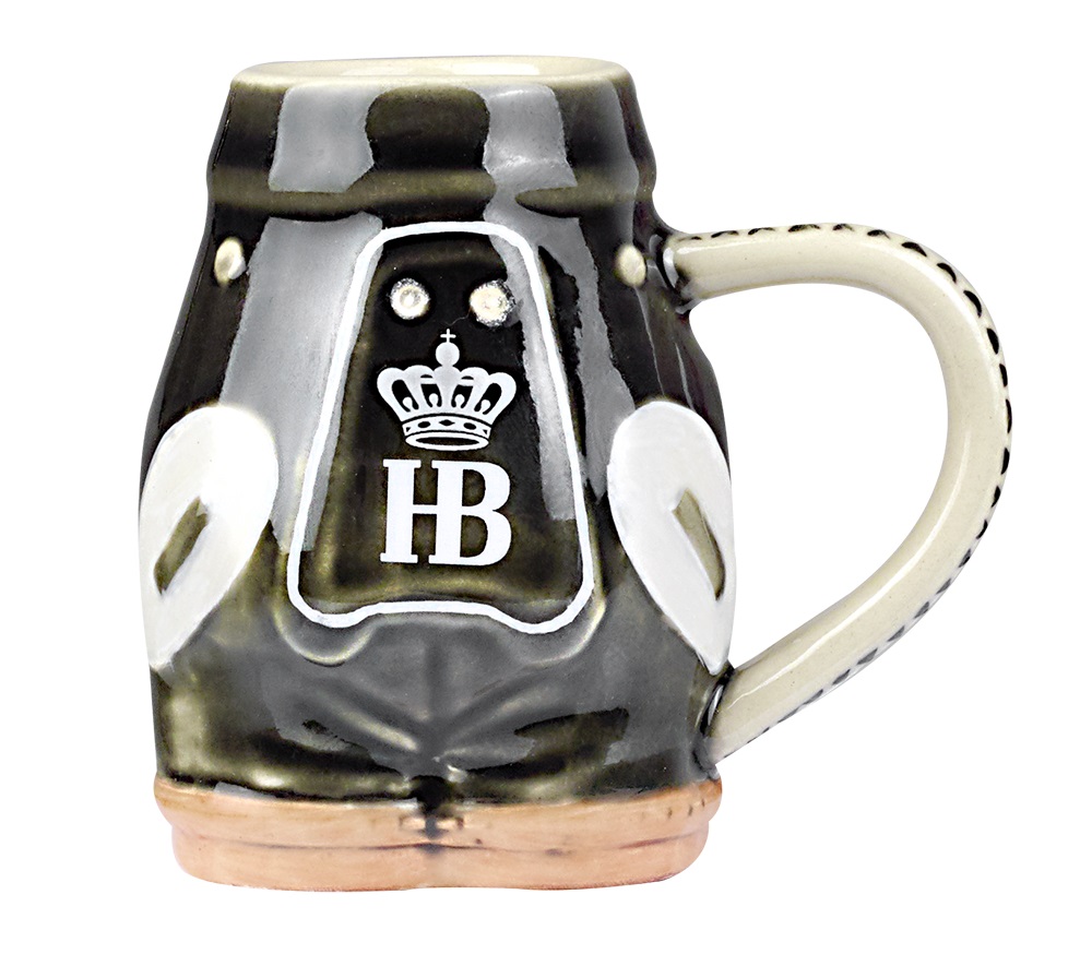 Steingut-Schnapsglas in Form einer Lederhose gestaltet. Das Glas ist bunt bemalt und zeigt eine braune Lederhose. Auf der Brust befindet sich das HB-Logo und einer Krone zu sehen. Seitlich befindet sich ein Henkel.