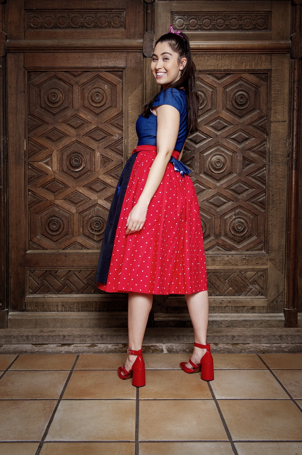 Frau in Dirndl Maibock von der Seite und leicht rückwärts: Blaues Oberteil, rote Schürze mit weißen Punkten, dunkelblaue Schürze darüber, rote Schuhe; Hintergrund mit geschnitzter Holztür.