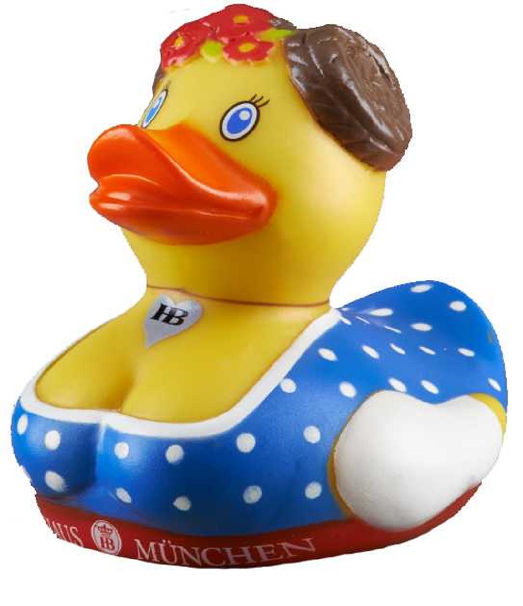 Gelbe Badeente im Dirndl-Design mit blauem Kleid und weißen Punkten, roten Lippen, braunen Zöpfen, Blumenschmuck im Haar und Herzanhänger mit 'HB'-Logo, seitlich links fotografiert.